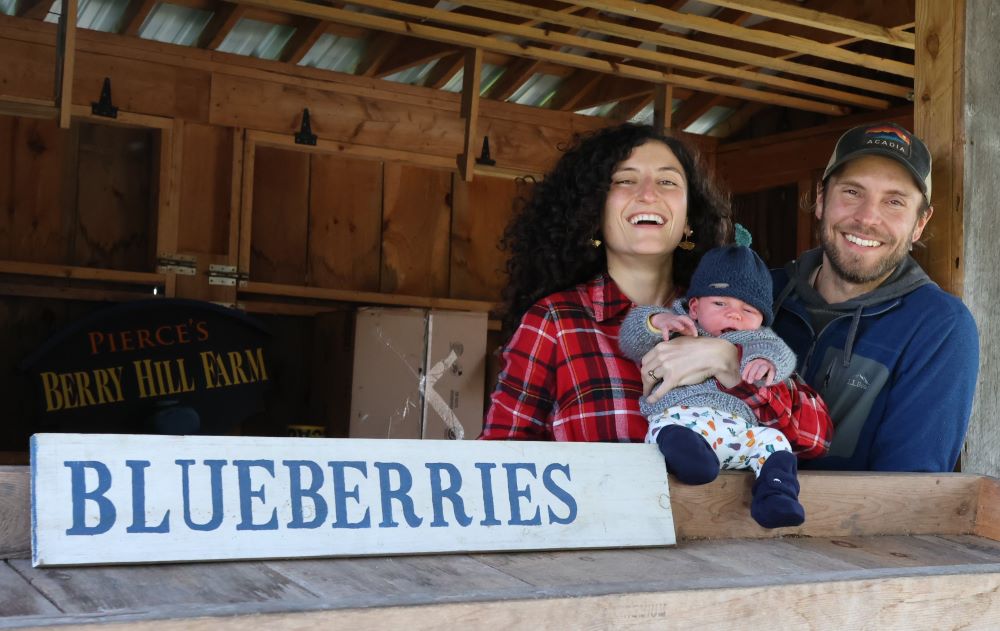 smiling woman and man hold baby behind blueberries sign