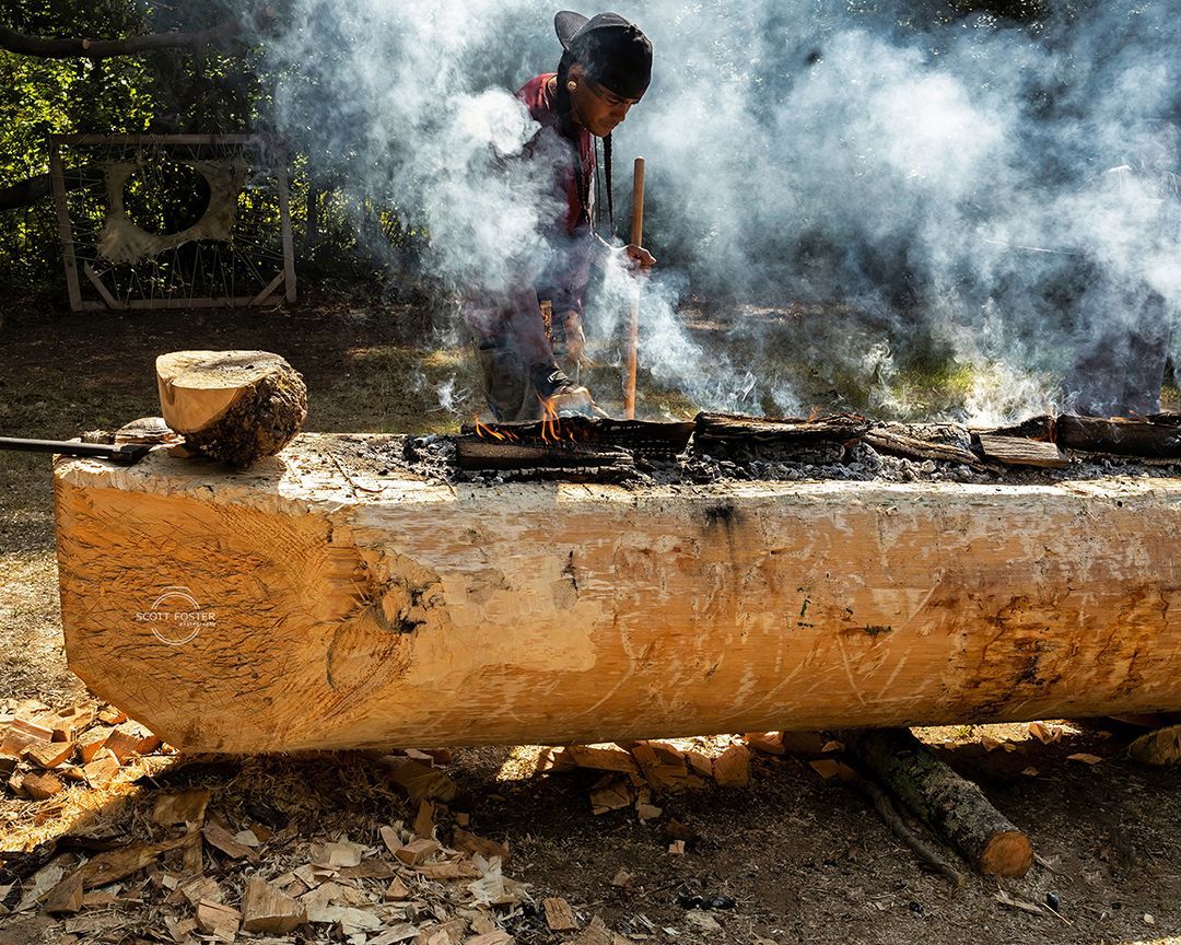 young man in braids and hat feeds fire in log canoe